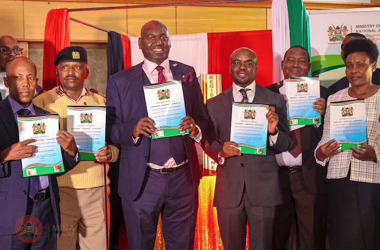 PSs Julius Bitok (Immigration and Citizen Services) and Raymond Omollo (Interior and National Administration) and other government officials display copies of the new guidelines on the issuance of IDs at the Kenya School of Government, Kabete