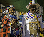 Proud parents Patricia and Thabo Msiza in front of the homestead. Patricia wears a red blanket associated with the initiation of her son, and the Long Tears (the long beaded strips that run from her headdress down to the ground) that signify the loss of her son but the gaining of a man. Thabo wears his family's totem (the animal skin decoration hanging over his chest) and carries his stick, and a more modern white shirt and matching hat painted with Ndebele designs.