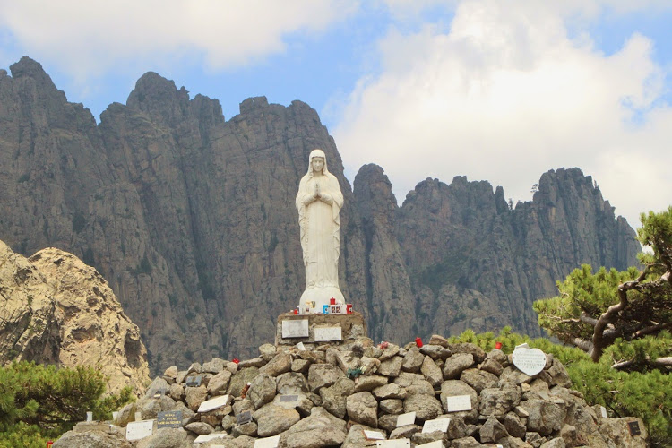 A statue of Our Lady of the Snows stands next to the road at the Col de Bavella mountain pass and is framed by the needle-shaped granite of the Aiguilles de Bavella in Corsica