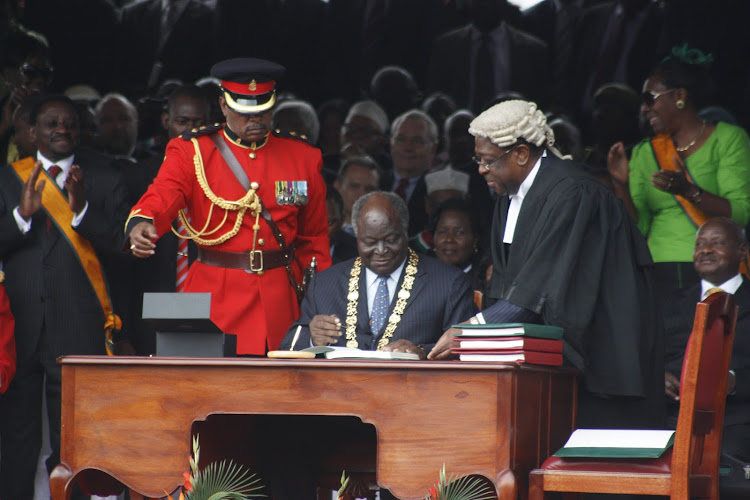 Former President Mwai Kibaki signs the documents of the new constitution as former AG Amos Wako looks on at Uhuru Park during the promulgation of the constitution ceremony in 2010.