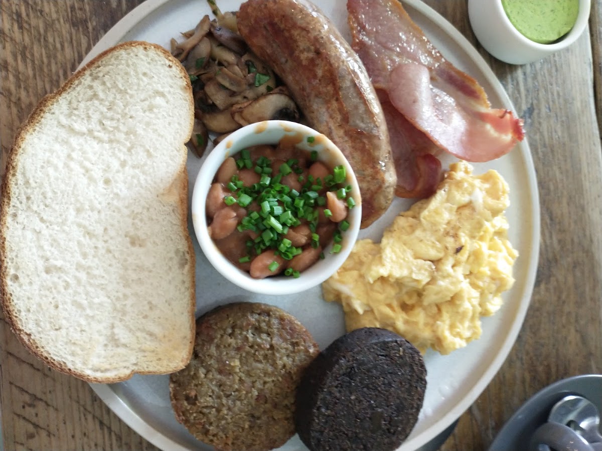 Gluten free butcher's breakfast, including gf sausage, black pudding, white pudding, beans, mushrooms, bacon, eggs, and gf sourdough.