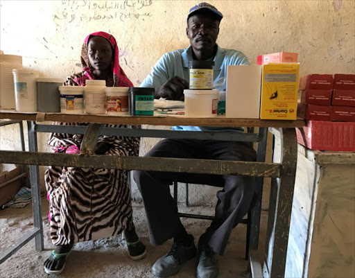 Mukuma Hamad, a volunteer health worker (L), and James Atai, a nurse, sit at a table displaying almost the total stock of basic medicines in the only health clinic in Hadara village, rebel-controlled Southern Kordofan. (© Skye Wheeler 2016)