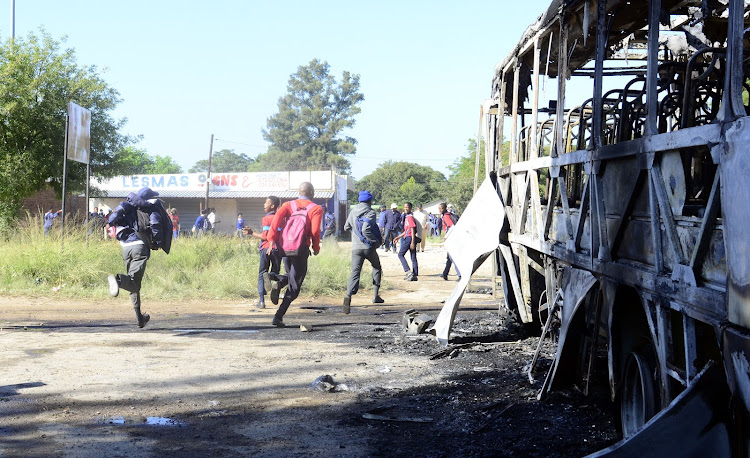 School children run near a burnt bus as residents of Mahikeng protested on the streets of Mahikeng demanding the resignation of Premier Supra Mahumapelo.