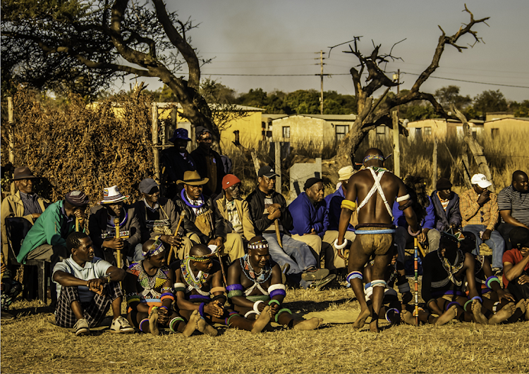 At the gifts ceremony, the initiates sit in a line on a grass mat with their fathers behind them with the elder men. At the opposite end of the field sit the mothers, mostly in their red blankets and mlinga koba (long tears). The seclusion boma can be seen behind on the left.