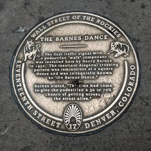 Sidewalk plaque marking the creation by traffic engineer Henry Barnes of the first traffic lights that included a pedestrian “Walk” signal. The arrangement triggered diagonal crossings that conjured ...