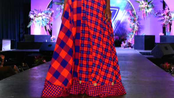 A woman wears a maxi, skater dress with red and blue Maasai pattern. The neck has red sheers and blue plain material across the arms.