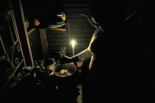 A woman prepares a meal using a primus stove by candlelight due to power cuts. The writer says the nation should look no further that the ANC's mismanagement of the entire state machinery for the genesis of problems such as power cuts, lack of safety and security and a failing education system.