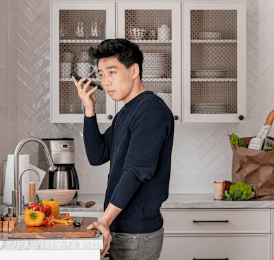 An Asian-American man stands at a kitchen counter holding his Android phone, listening to a screen reader verbalising a recipe.