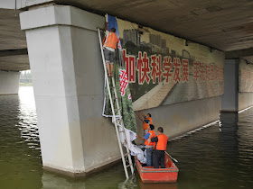 man on ladder and men in boat covering the slogan "加快科学发展 率先转型跨越" with another sign