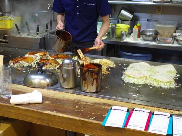 鉄板の上で沢山お好み焼きが作られてる。ソースを塗ってるところ。