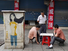 two men playing xiangqi next to an electric box with a drawing of a woman