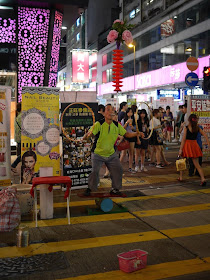 street performer balancing on a small barrel while twirling hoops and balancing a flower and saucers with a curved rod from his mouth in Hong Kong