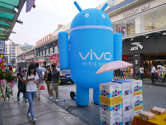 a large blue inflated Android robot with the Vivo logo on a sidewalk at a Zhuhai shopping district