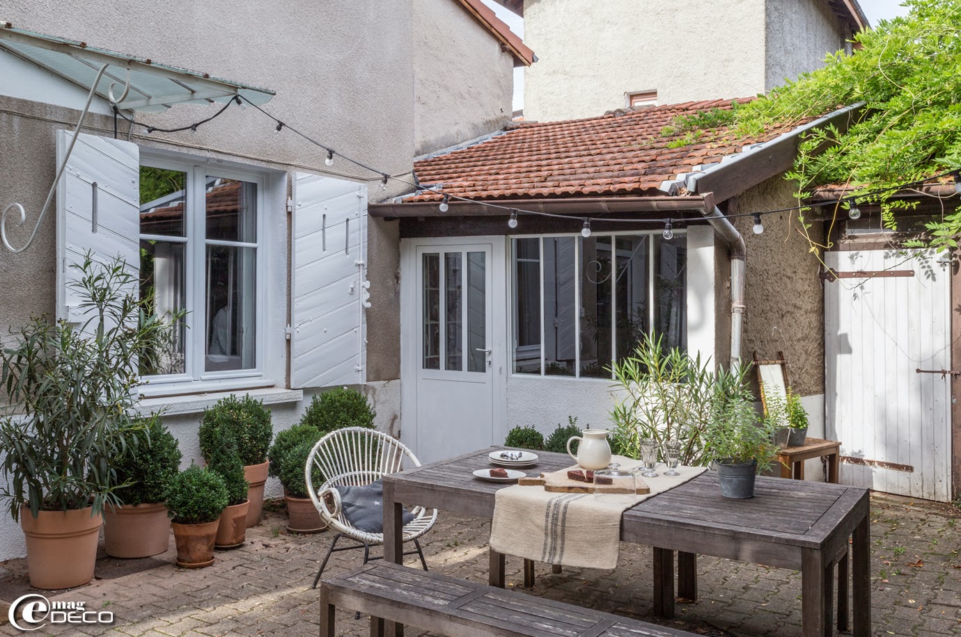 Chez Florence Bouvier, la terrasse devant la maison est éclairée par une guirlande lumineuse 'Un Esprit en Plus'. Fauteuil en rotin chiné aux Puces du Canal à Villeurbanne et sac à grain posé en chemin de table chiné à Avignon