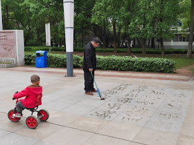 child on tricycle rides by a man doing water calligraphy