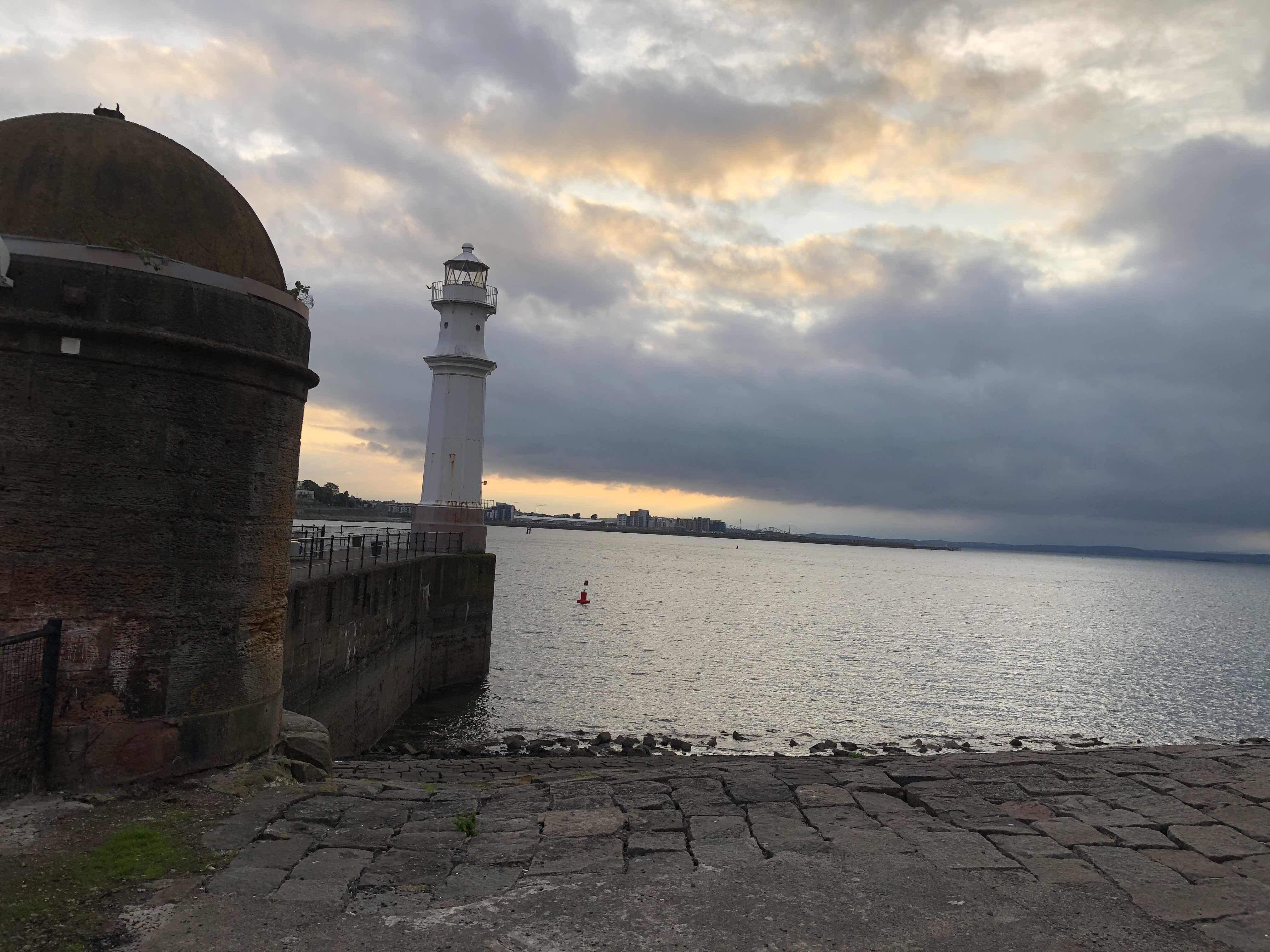 A slender lighthouse almost hidden by a large brick object in the foreground of the image. In the background is the Firth of Forth, the wide river estuary to the north of Edinburgh. 