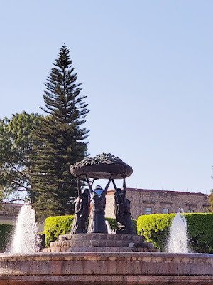 Bronze statue of thee native Mexican women at the center of a fountain. They are unified by holding up a giant woven platter of fruit. Locals have added cloth masks and scarves to the figures.