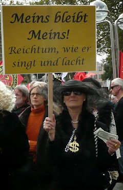 Aufgetakelte Spaß-Demonstrantin mit goldenem Dollarschmuck und Banknoten in den Händen, Transparent: »Meins bleibt Meins! Reichtum, wie er singt und lacht«.