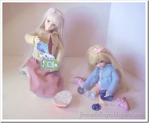 Two ball jointed dolls assembling a Kinder Joy toy.  The larger doll is trying to read the instructions and the smaller younger doll is putting the toy together with no effort at all.