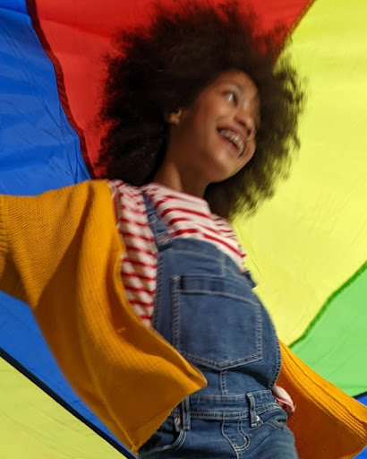 A younger child wearing overalls and an orange sweater smiles while playing under a colorful parachute. Details of their face and bright smile come into focus against a blurred background of color.