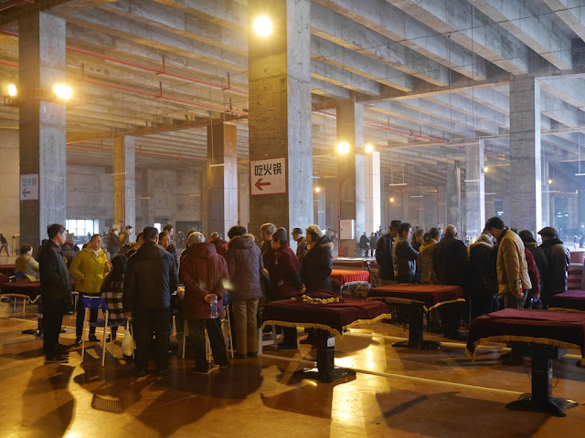 people standing while playing mahjong in the large indoor area like a large parking lot