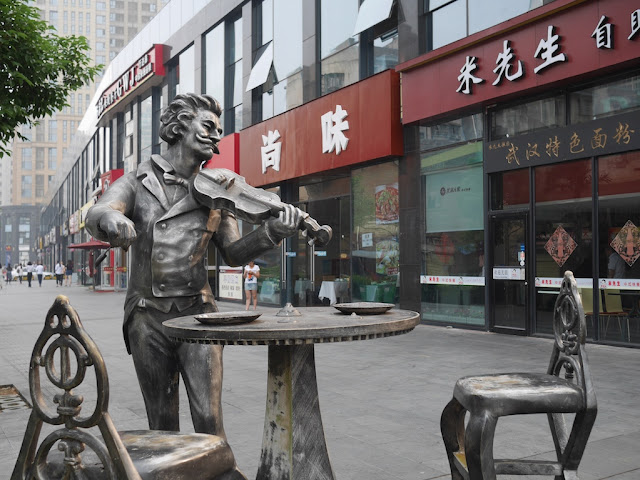 statue of a man playing a violin next to a table and chairs behind Kaifu Wanda Plaza