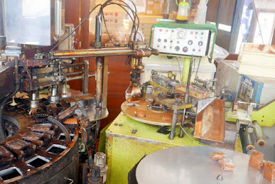 Automated ningyo yaki machine. The crane on the right side carries the cooked cake to the machine on this right side to cool and get packaged down the little slide of plastic you see. On the left you can see the brushing inside the mold and then the squeezing of dough to the left of that