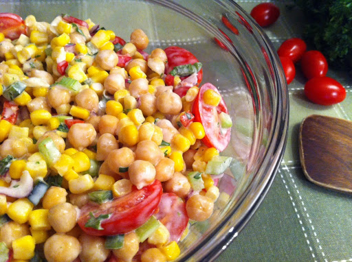 A glass bowl with a mixture of chickpeas, corns, and tomatoes on a green table mat with a wooden spoon and whole cherry tomatoes.