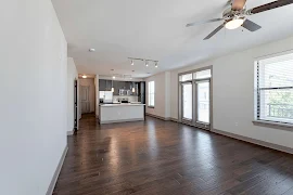 Living room with dark wood-inspired floors, neutral trim, light walls with french doors to balcony & the kitchen in the back.