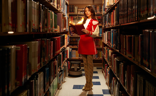 Madison Beer in a red varsity jacket and red skirt in a library holding a book that is on fire.