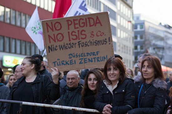 Demonstrantinnen, Fahnen, Plakat: »No Pegida, no Isis & Co. Nie wieder Faschismus! Düsseldorf, Türkei Zentrum.«.