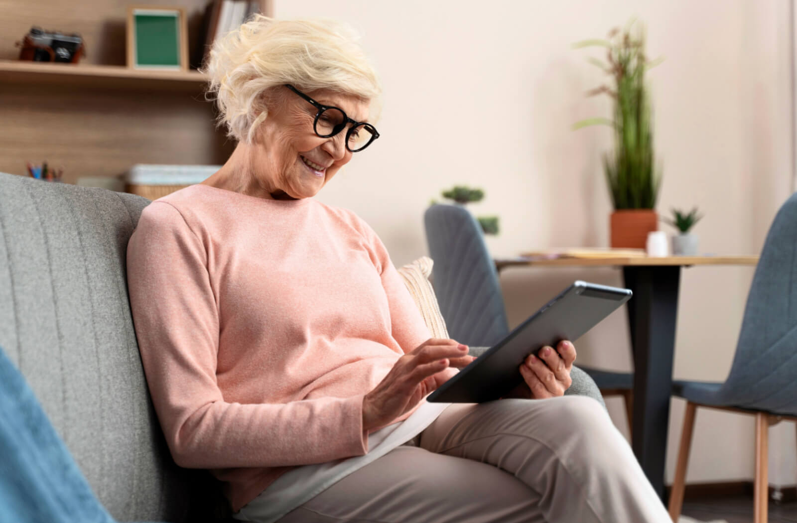 A senior woman wearing black glasses sitting on a couch and using a tablet while smiling.
