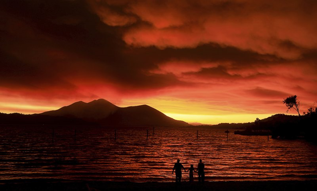 Evacuees from Lucerne, from left, Ken Bennett with Ember Reynolds, 8, and Lisa Reynolds watch the sunset as smoke from the Ranch Fire rises into the sky at Austin Park Beach in California’s Clearlake, with Mount Konocti in the background, 6 August 2018. Photo: Kent Porter / The Press Democrat / AP