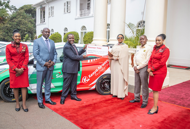 Kenya Airways Corporate Executive Officer Allan Kilavuka hands a dummy ticket to SPorts CS Amina Mohamed as Sports PS Joe Okudo (2nd L) and Safari Rally Project CEO Phineas Kimath (2nd R) look on.