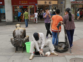 man sitting on a sculpture's small stool while attending to two puppies; other people have their photograph taken with the scullpture