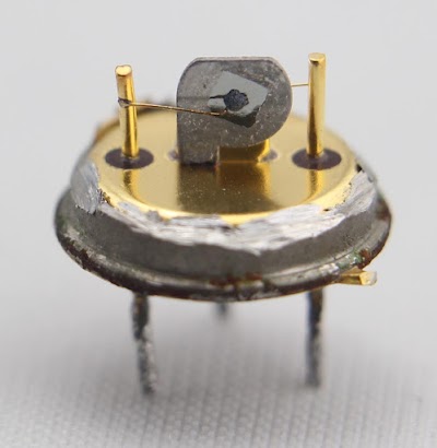 Inside a failed IBM 083 germanium transistor. The silver-colored square in the middle is the germanium die, wired to the base pin. The dot in the middle is tin alloy, forming the collector, with a wire to the collector pin on the left. A smaller dot on the other side of the germanium die forms the emitter, wired to the pin on the right.