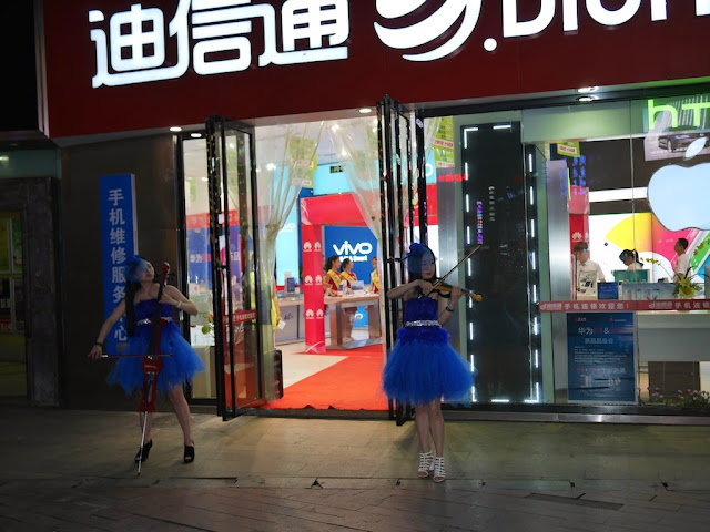 two young women in blue dresses playing electric string instruments outside a mobile phone store in Changsha