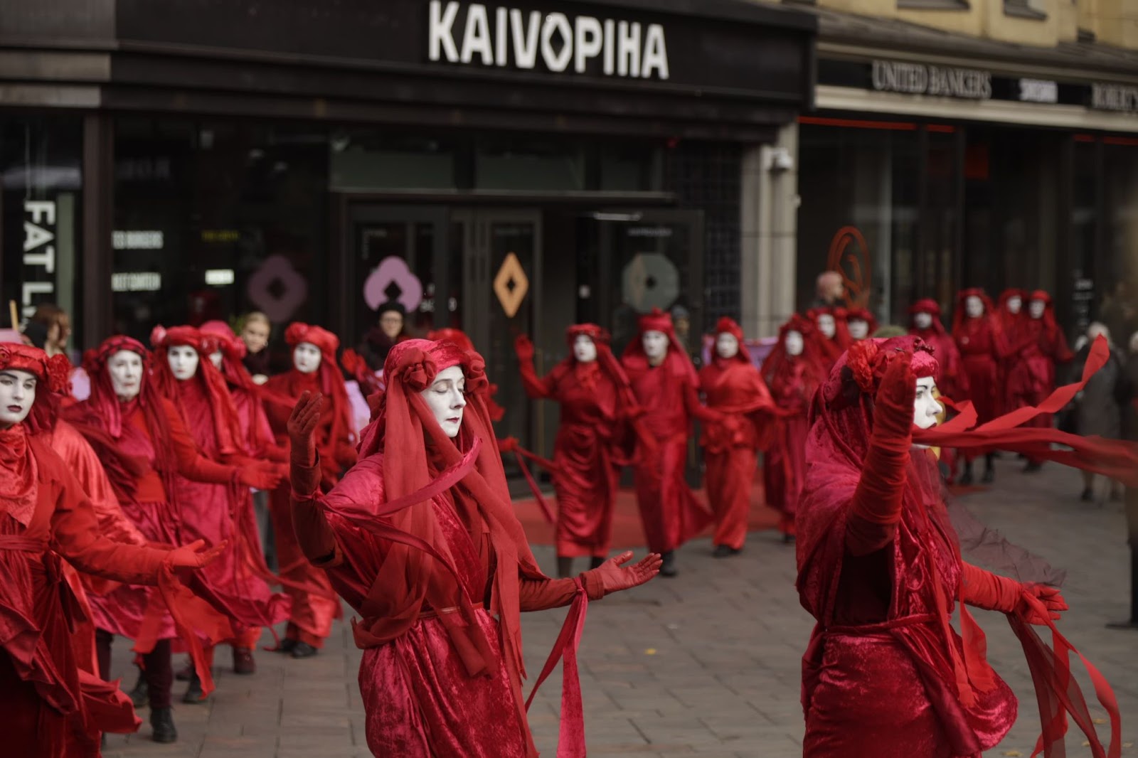 Red brigade rebels dance in a line through a shopping mall, they faces calm and thier red ribbons seeming to float in the air.