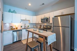 Modern apartment kitchen with white cabinets, stainless steel appliances, and a wooden dining table with stools.