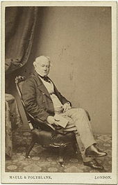 A sepia cabinet photograph of a middle-aged man relaxing in an armchair. He is dressed in mid-nineteenth century clothing and holds a book in one hand.