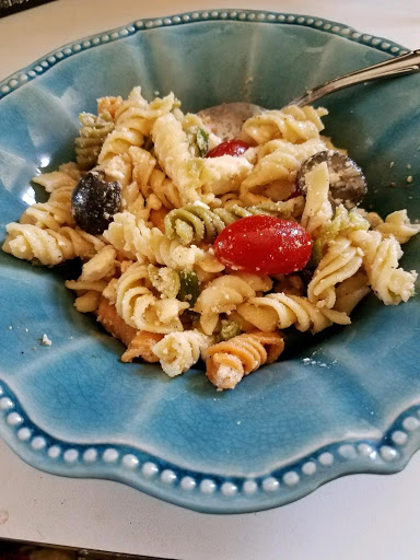 Tri-colored rotini pasta with sliced black olives, halved cherry tomatoes, spring onions, feta cheese with parmesan and asiago, with a fork in a blue bowl