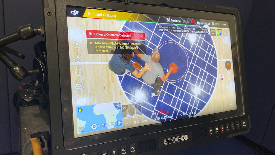 Close up image of a drone camera monitor screen showing a few technical tools and the image that the camera is capturing of two people playing basketball on an indoor court.