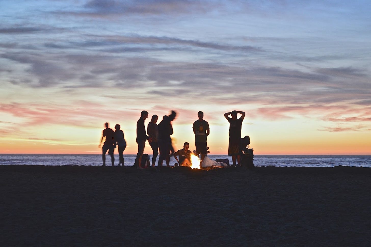 Several people silhouetted around a campfire on the beach at sunset.