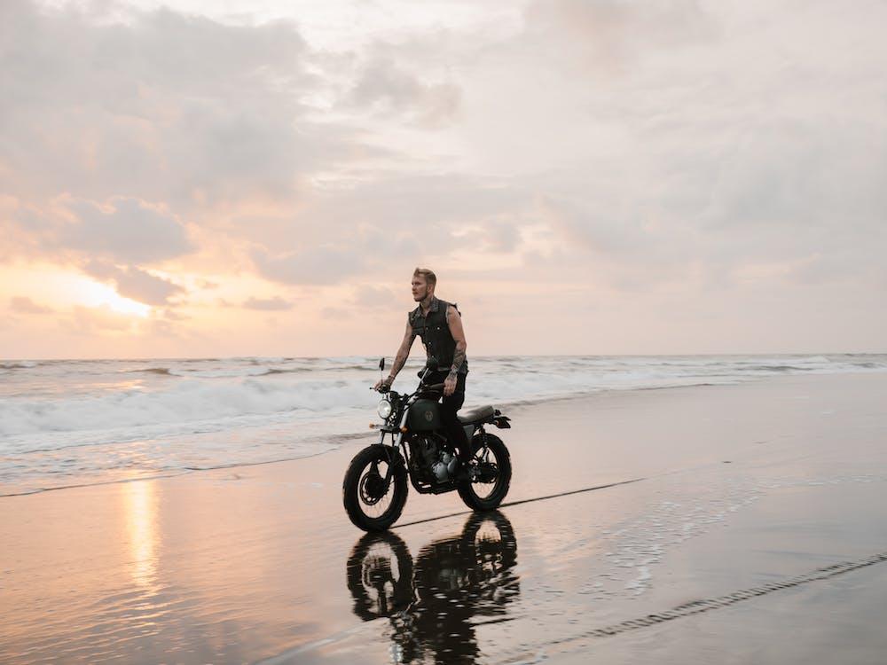 Free Tattooed biker riding modern motorcycle reflecting on sand of ocean beach near foamy waves under picturesque cloudy sky at sundown Stock Photo