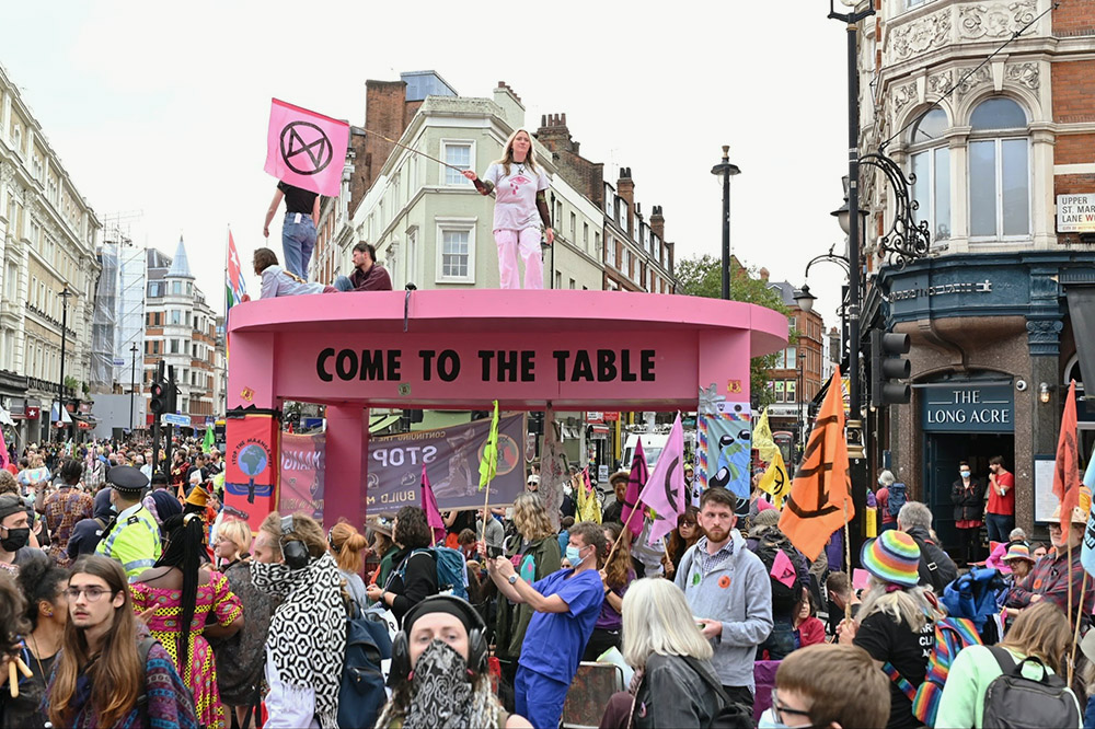 A giant pink table rises up from a crowd of people on a London crossroads.Two people stand on top of the table waving pink XR flags.