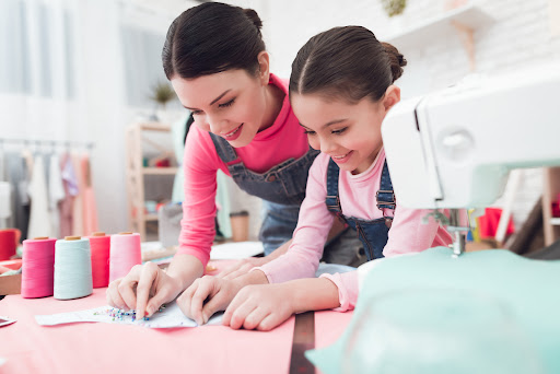 A little girl and woman together construct clothes. They are with Mom in the sewing workshop. They are in a good mood. They use pins.