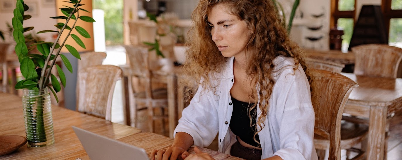 Woman on her laptop sitting at a table next to a vase with foliage.