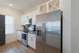 Black tile backsplash, stainless steel appliances, white cabinets, stone counters in kitchen, plank floor, window with blinds