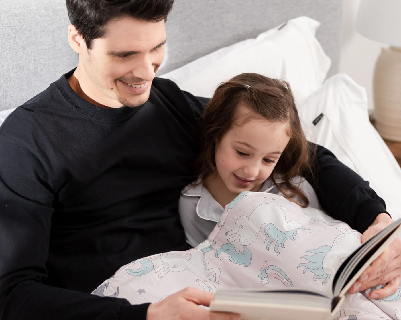 A man reading to his daughter in bed while under a Hush Kids Weighted Blanket in unicorn pink pattern.