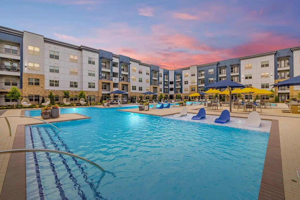 Community swimming pool with lounge furniture, sun shelf, sculpture, apartment buildings in background at dusk